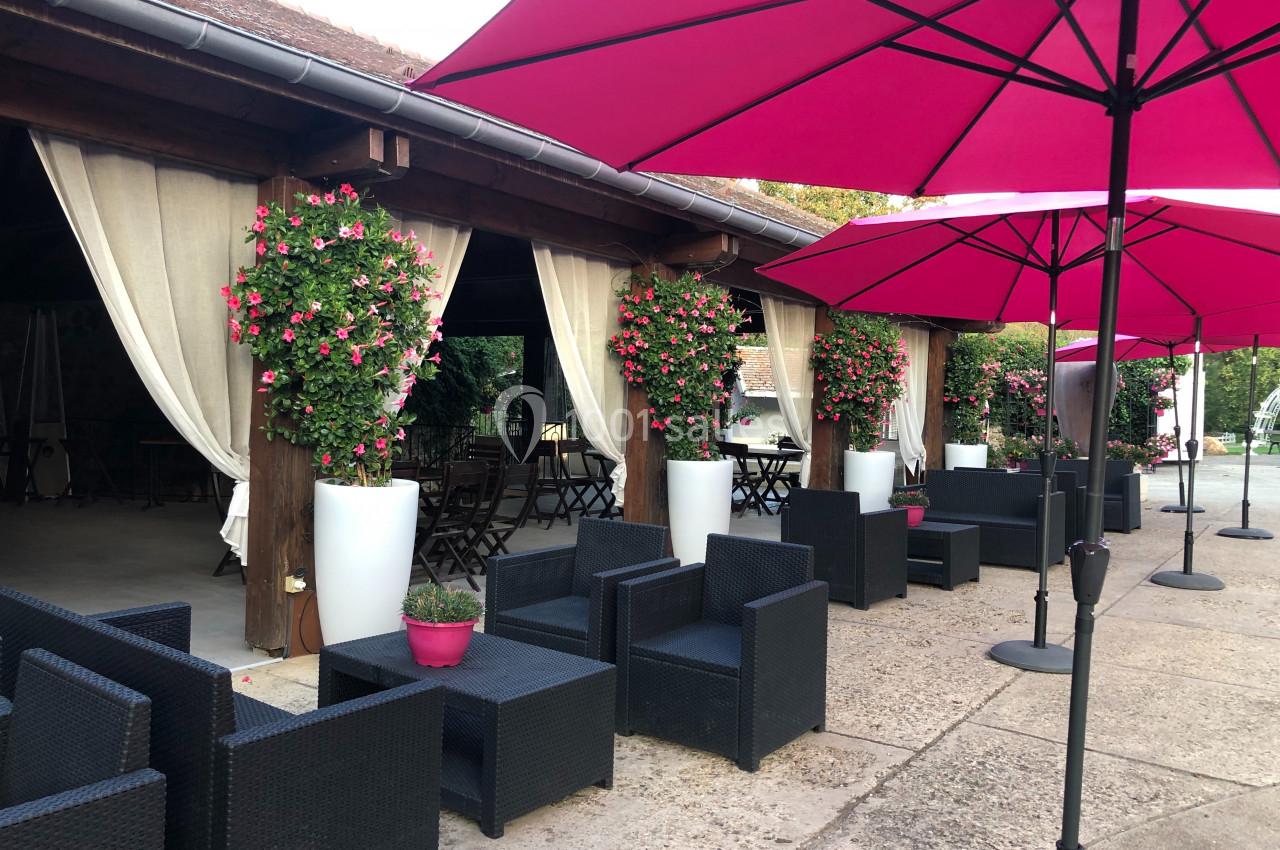 Terrasse aménagée avec des fauteuils en résine noire, des parasols roses et des plantes fleuries en pots blancs.