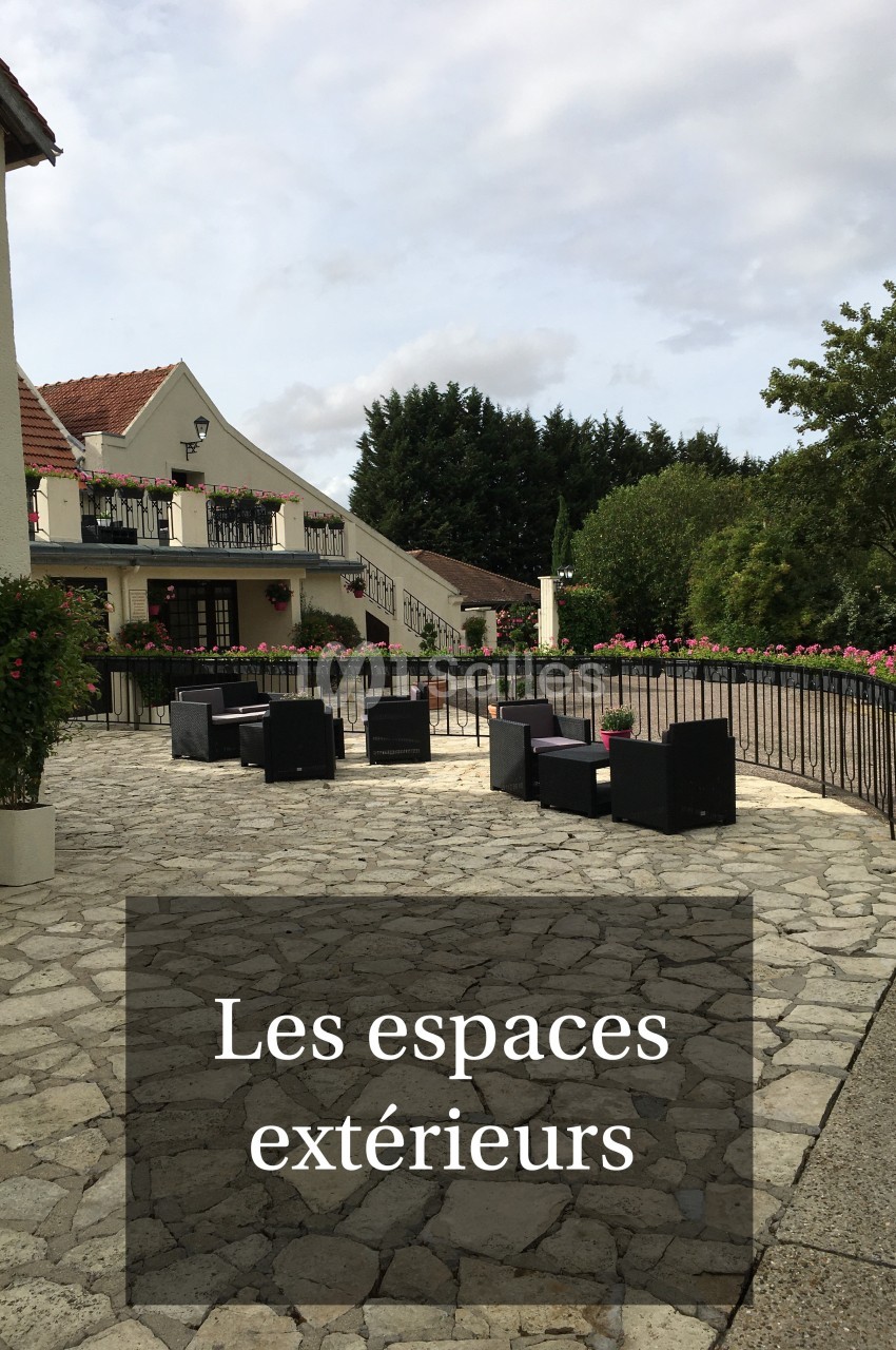 Terrasse en pierre avec fauteuils noirs, entourée de fleurs et d'arbres, devant un bâtiment à balcons fleuris.