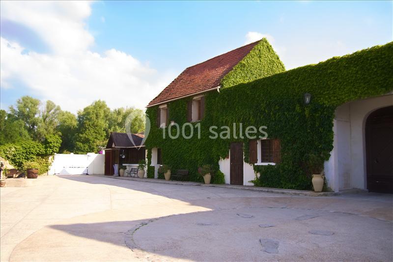 Maison en pierre recouverte de lierre avec une cour pavée, entourée de verdure et ciel dégagé.