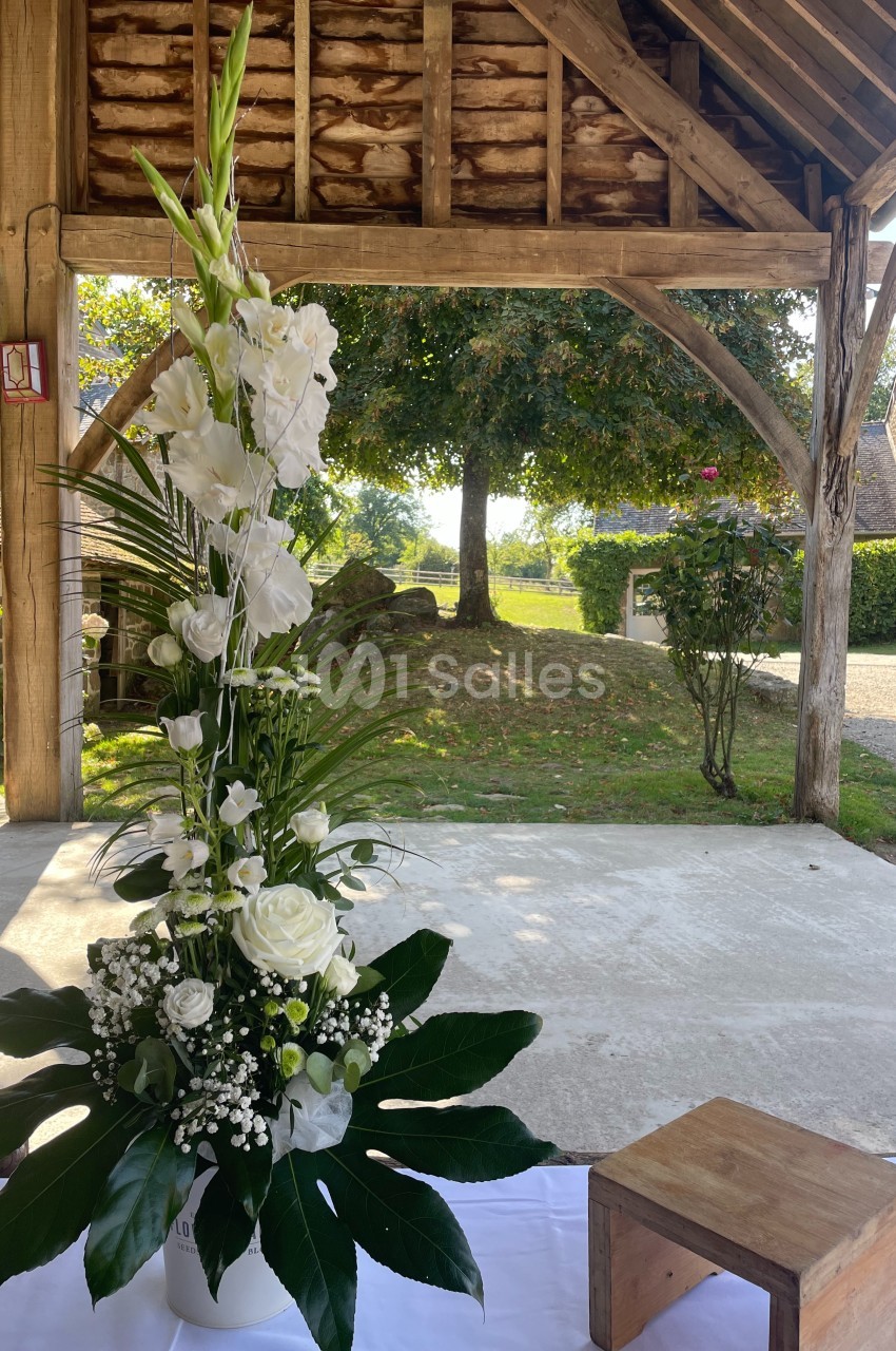 Composition florale blanche avec des roses et des glaïeuls, posée sur une table sous un abri en bois, vue sur un jardin.