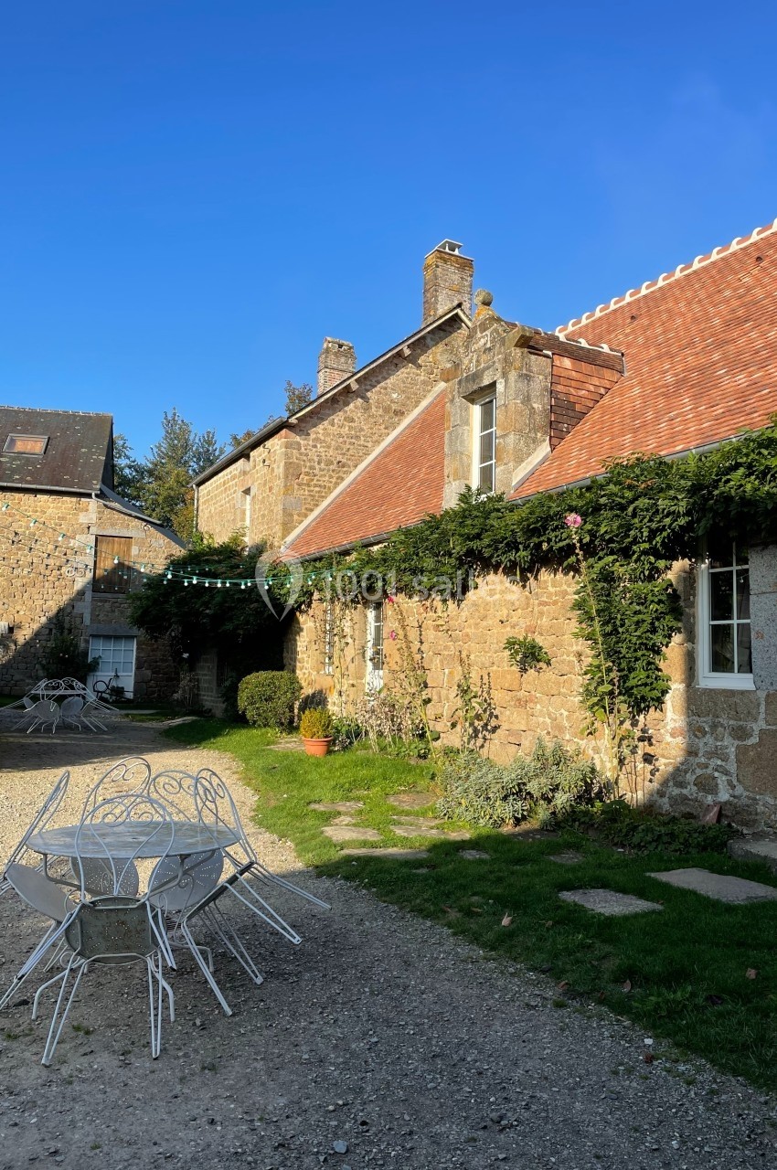 Cour d'une maison en pierre avec toit en tuiles, table et chaises blanches en métal, entourée de verdure.