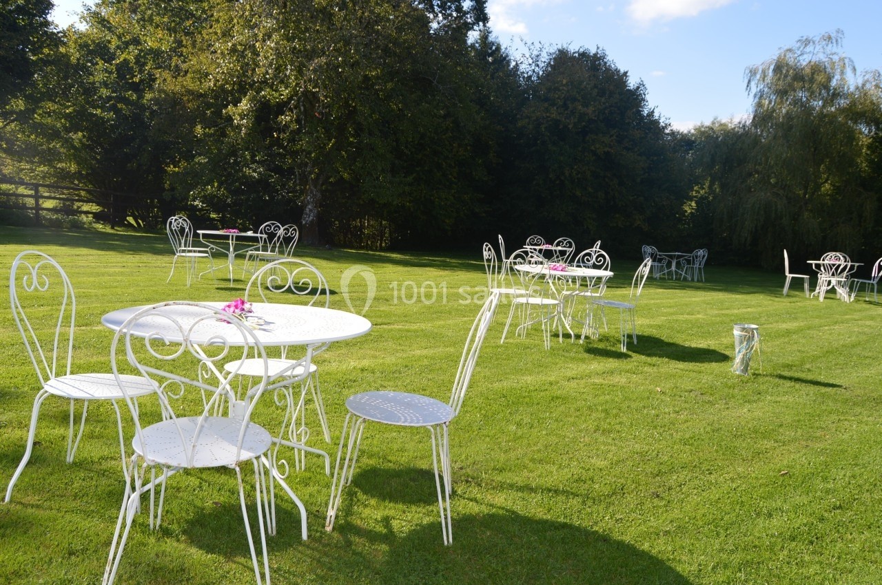 Tables et chaises blanches en métal disposées sur une pelouse verdoyante, entourées d'arbres.