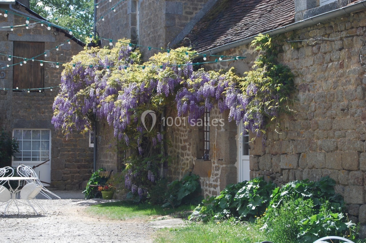 Façade en pierre d'une maison ancienne ornée de glycines en fleurs, avec une cour et des meubles de jardin.