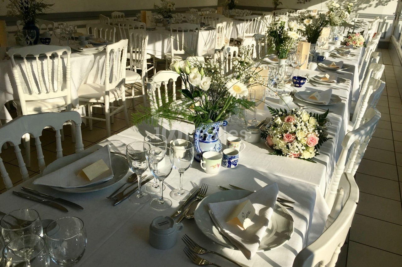 Tables de réception élégamment dressées avec nappes blanches, vaisselle, verres et bouquets de fleurs.