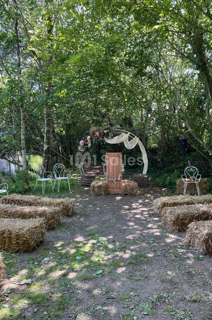 Espace extérieur aménagé pour une cérémonie, avec des bottes de paille, des chaises blanches et une arche décorée.