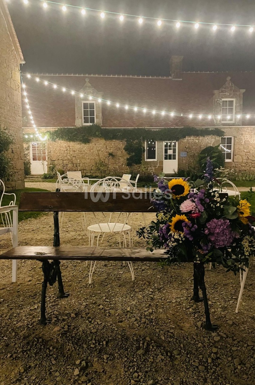 Banc en bois décoré de fleurs colorées, placé dans une cour en soirée éclairée par des guirlandes lumineuses.
