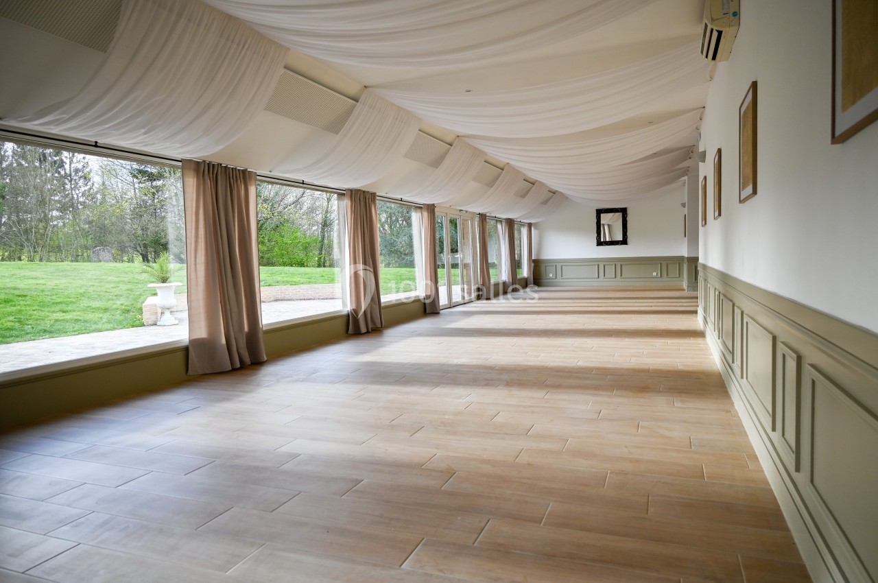 Salle lumineuse avec parquet en bois, grandes baies vitrées donnant sur un jardin verdoyant, plafond drapé de tissu blanc.