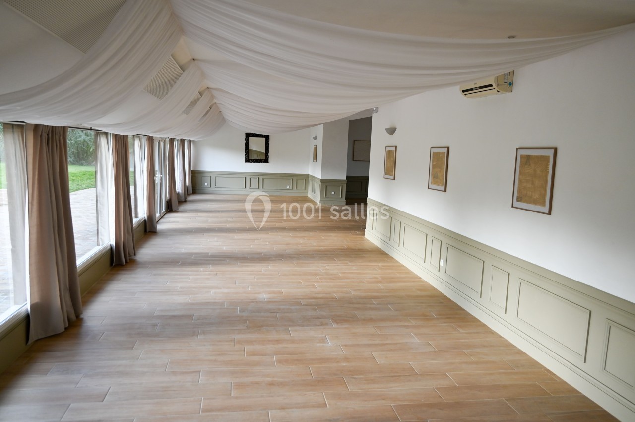 Salle lumineuse avec parquet en bois clair, murs blancs décorés de cadres et grandes fenêtres donnant sur l'extérieur.