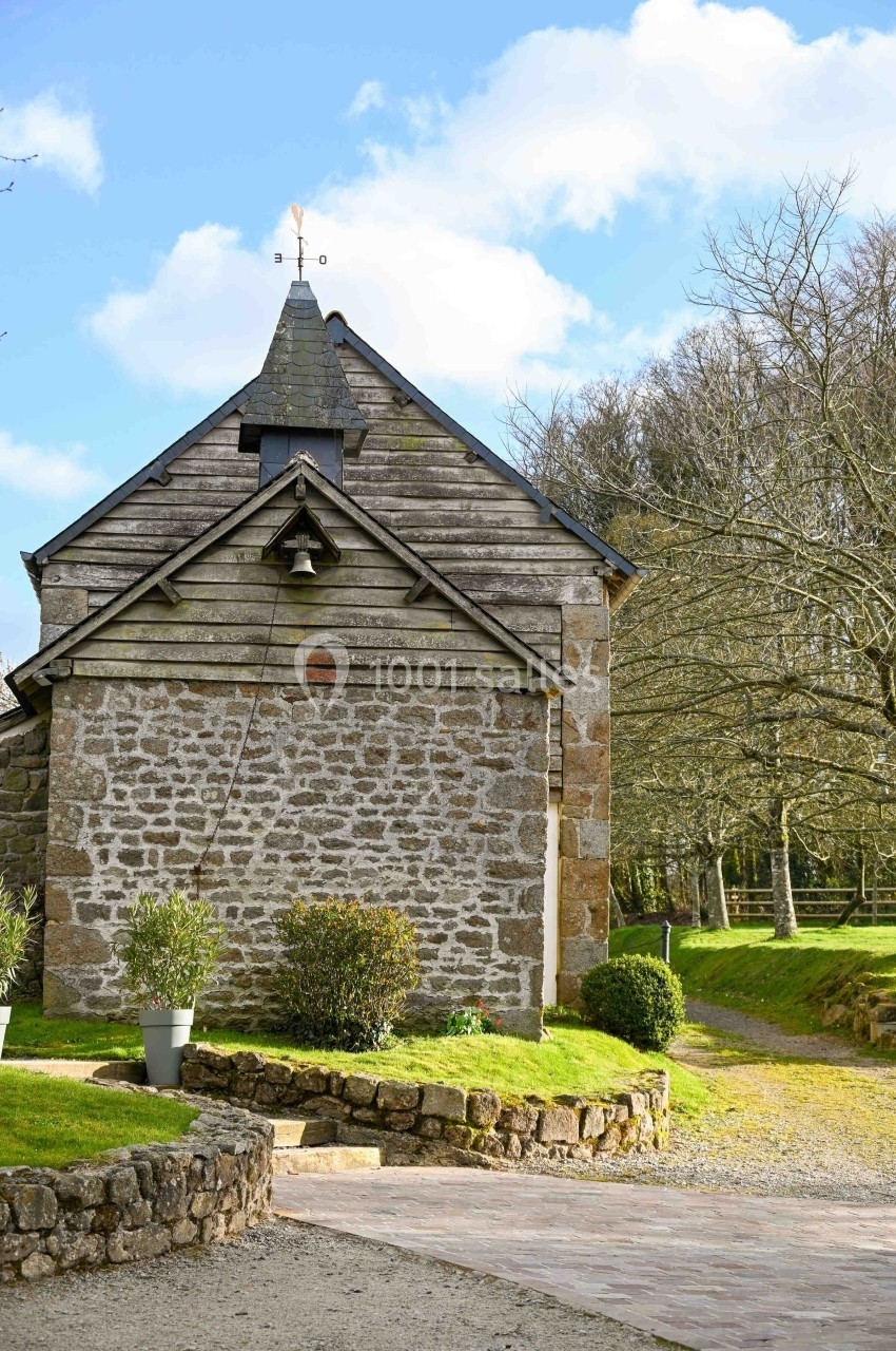 Petite bâtisse en pierre avec un toit en ardoise, entourée de végétation et d'un chemin pavé sous un ciel bleu.