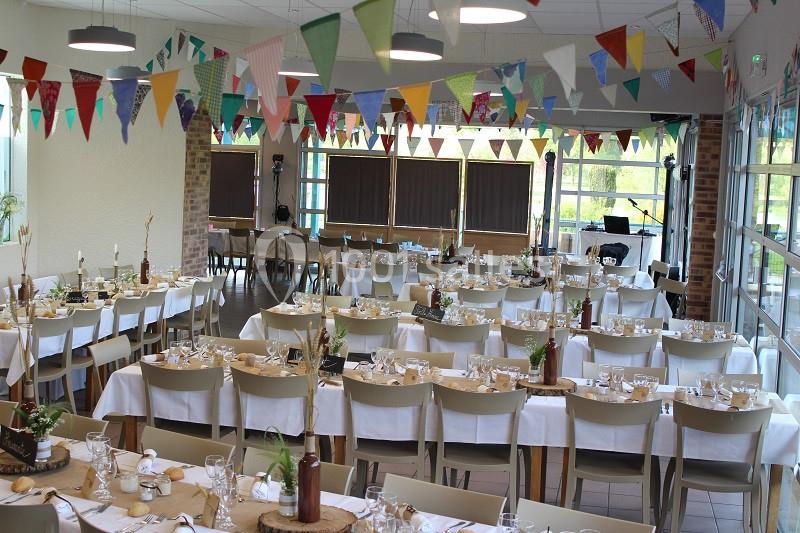 Salle décorée pour un événement avec des tables dressées, nappes blanches, guirlandes colorées et grandes baies vitrées.