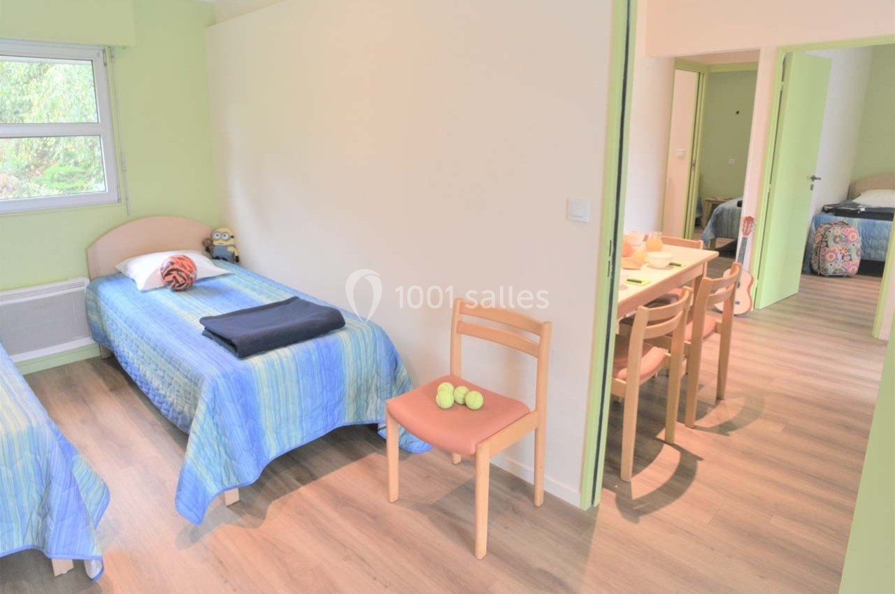Deux lits simples avec couvre-lits bleus dans une chambre lumineuse, vue sur une salle à manger adjacente.