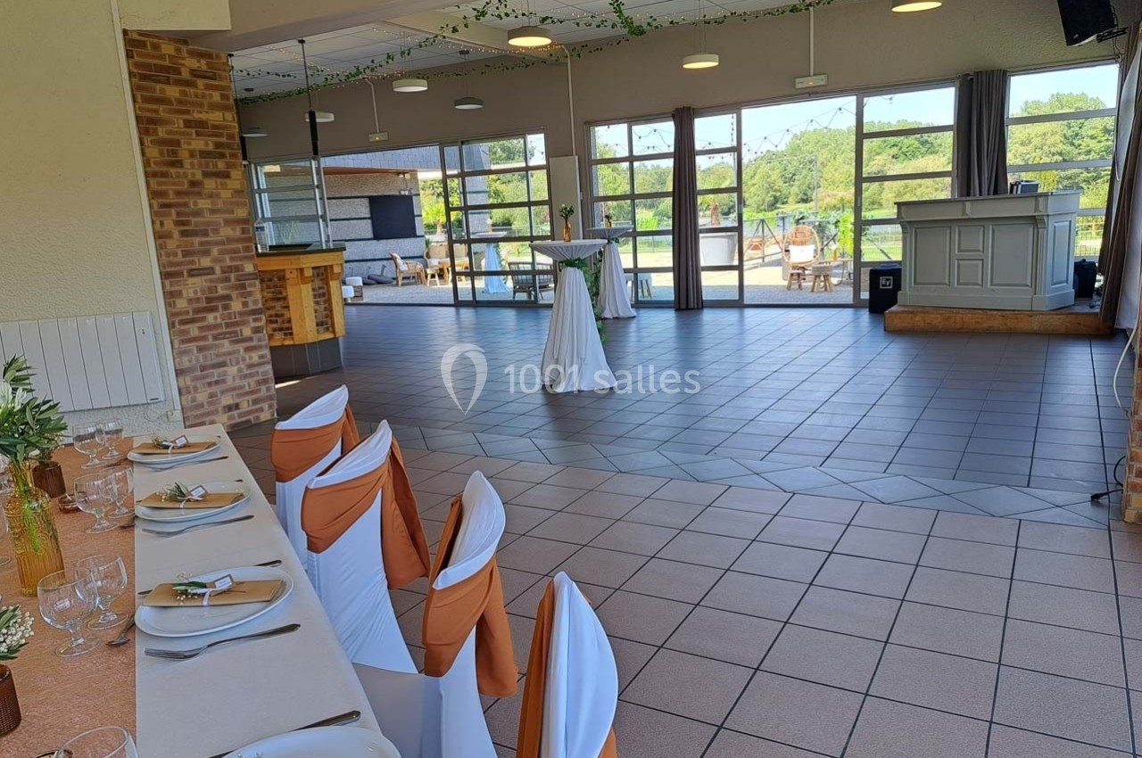 Salle lumineuse avec tables dressées, chaises décorées, bar et grandes baies vitrées donnant sur un espace extérieur…
