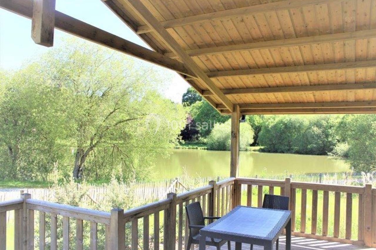 Terrasse en bois couverte avec table et chaises, donnant sur un étang entouré de végétation verdoyante.