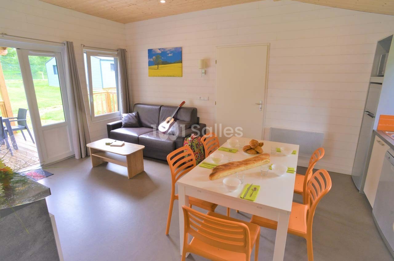 Salon lumineux avec canapé, table à manger, chaises orange, guitare posée, et vue sur terrasse et jardin.
