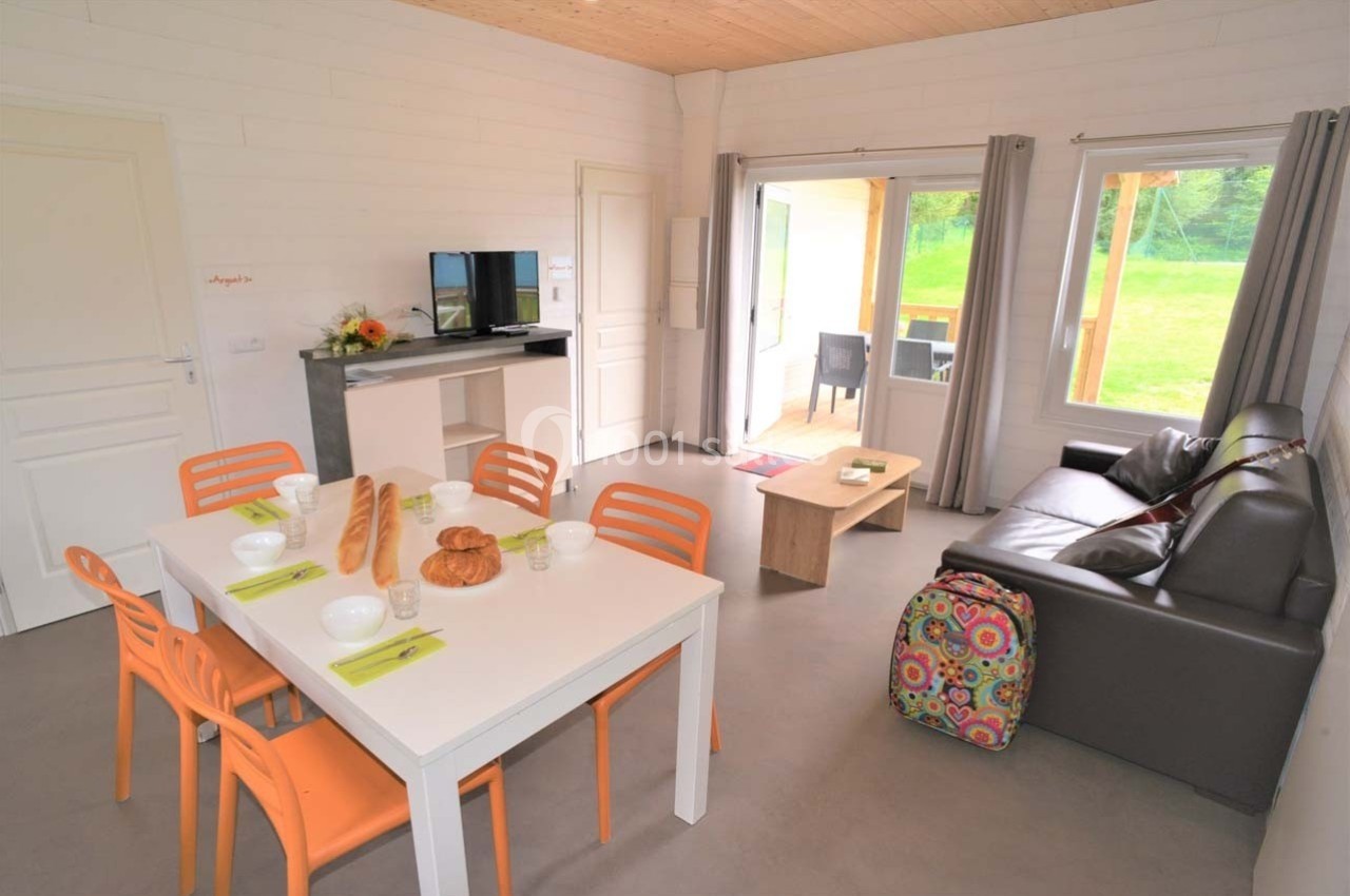 Salle de séjour lumineuse avec table à manger, canapé, télévision, et vue sur un jardin à travers une baie vitrée.