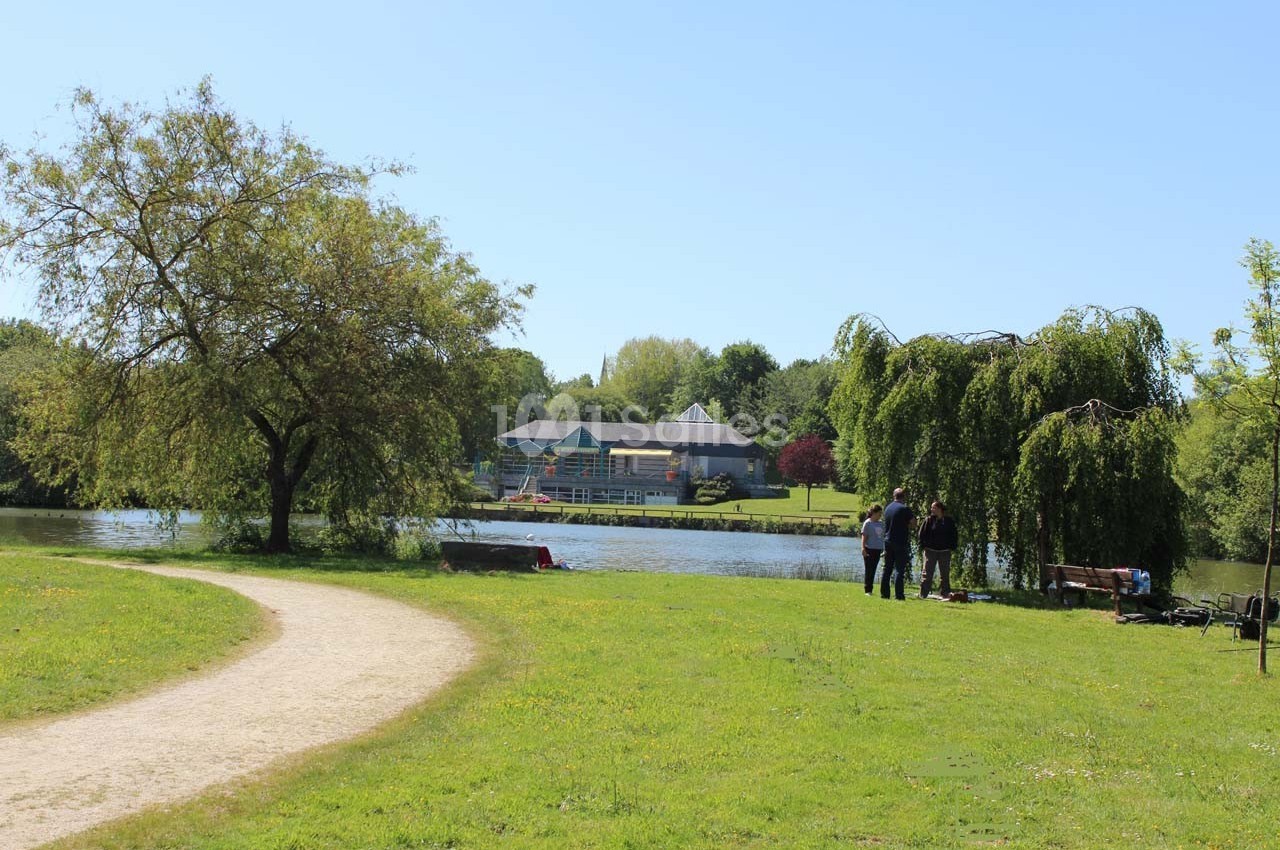 Un sentier courbé longe un parc verdoyant avec un lac, des arbres et des bâtiments en arrière-plan.