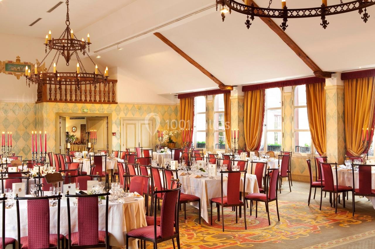 Location salle Gressy (Seine-et-Marne) - Le Manoir de Gressy #2 Salle de réception élégante avec tables dressées, chaises rouges, lustres en fer forgé et grandes fenêtres avec rideaux…