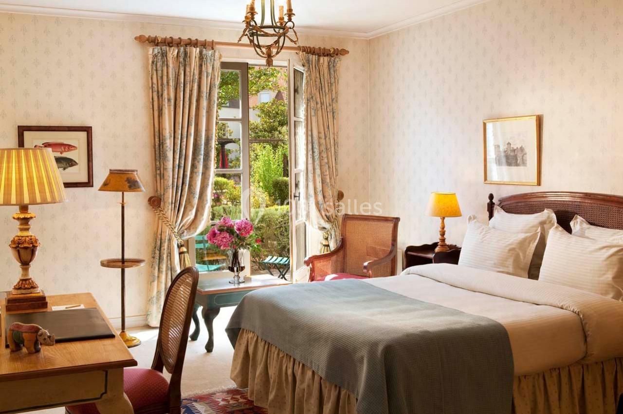 Location salle Gressy (Seine-et-Marne) - Le Manoir de Gressy #26 Chambre lumineuse avec lit double, bureau en bois, fauteuils et vue sur un jardin à travers une porte-fenêtre ouverte.