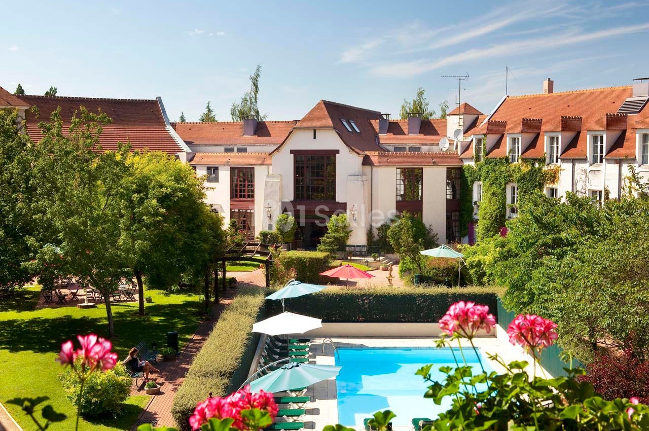 Vue d'un hôtel avec piscine extérieure, parasols, jardins verdoyants et bâtiments aux toits en tuiles rouges.