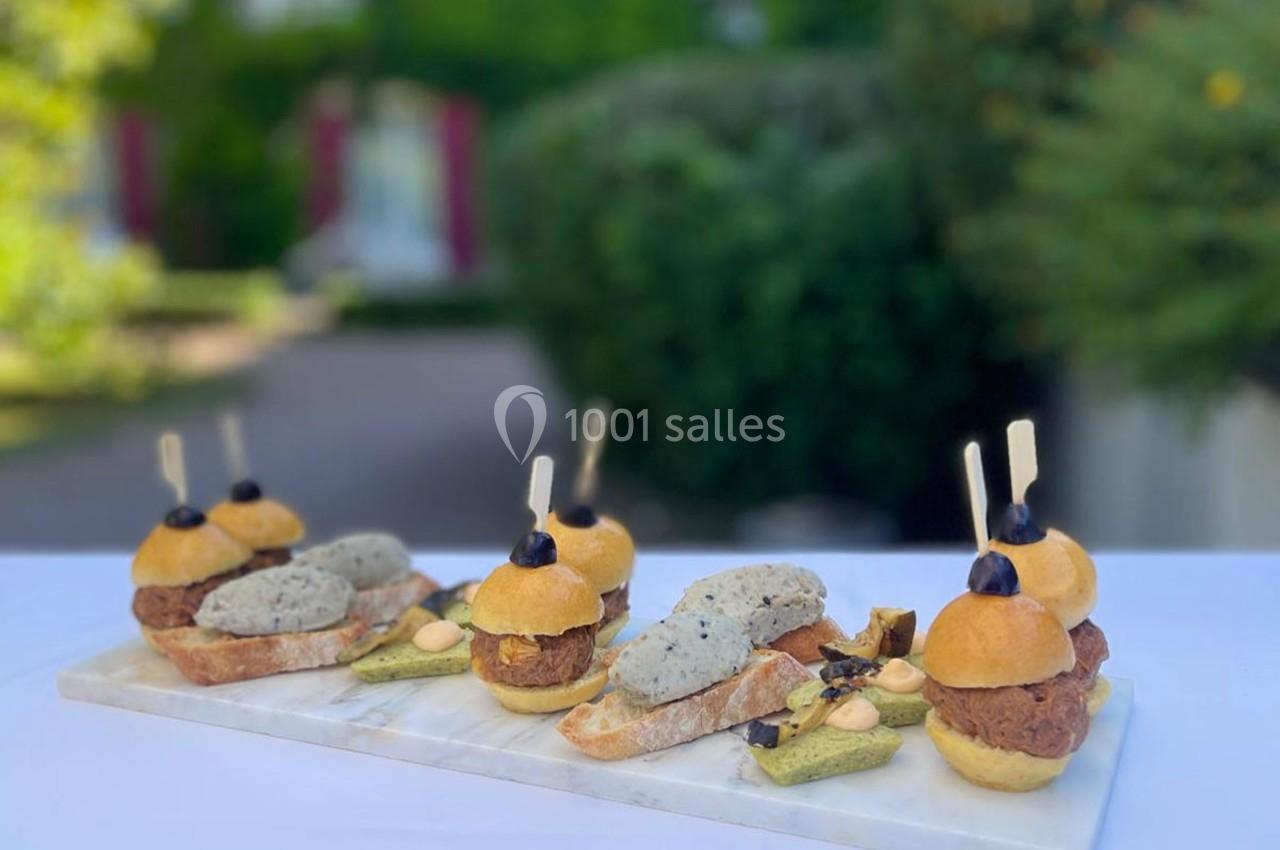 Location salle Gressy (Seine-et-Marne) - Le Manoir de Gressy #18 Plateau de mini-burgers et amuse-bouches variés présentés sur une table en extérieur, avec un jardin en arrière-plan.