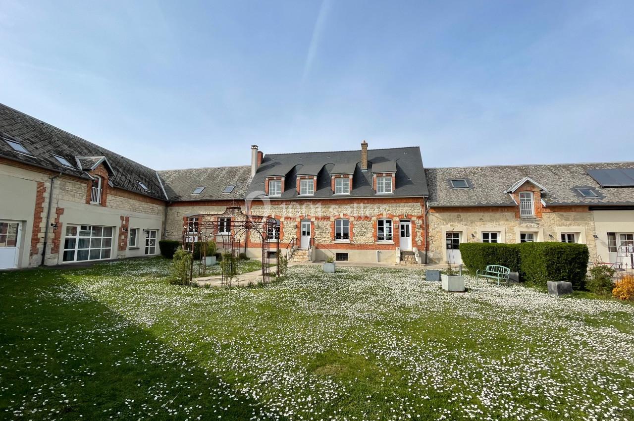 Grande maison en pierre avec toit en ardoise, entourée d'une cour herbeuse parsemée de petites fleurs blanches.