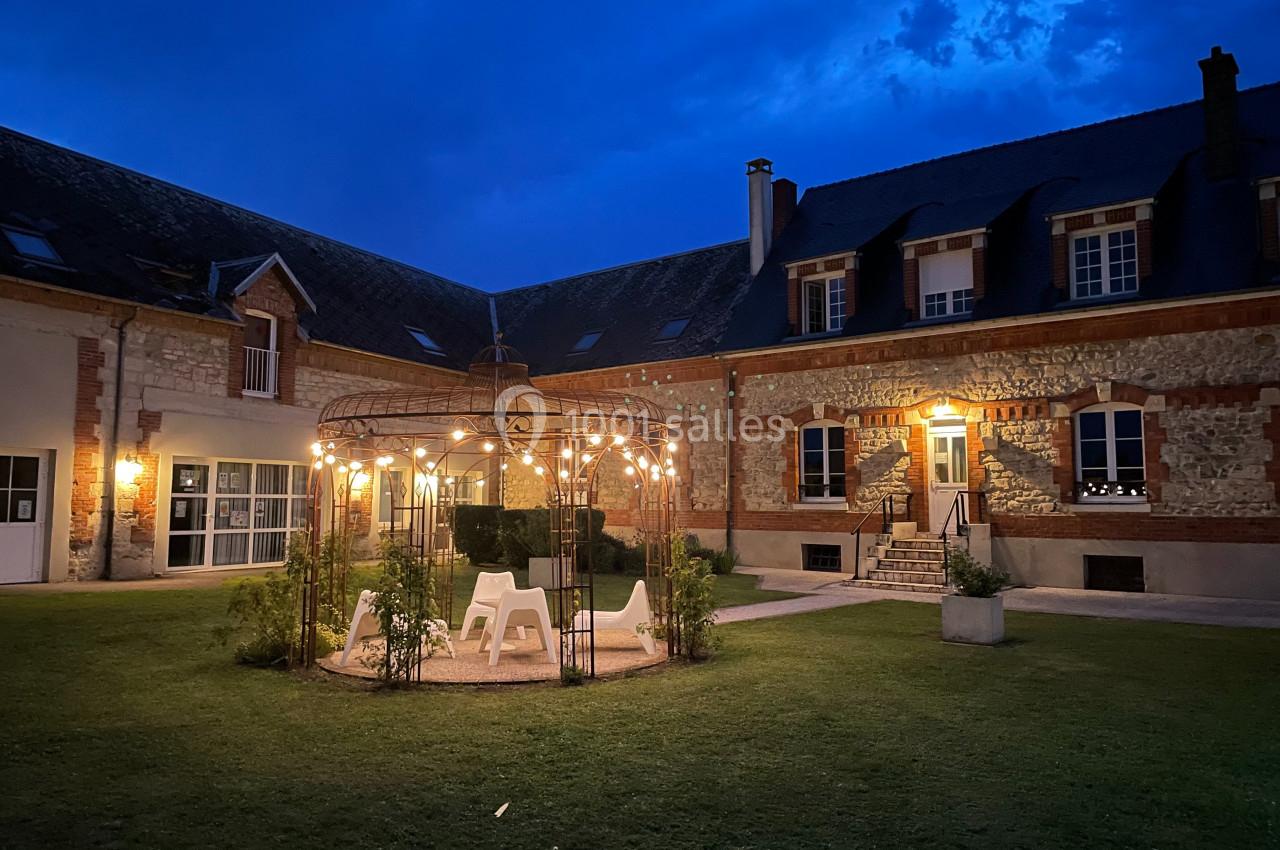 Cour intérieure illuminée en soirée, avec une pergola décorée de guirlandes lumineuses et des bâtiments en pierre.