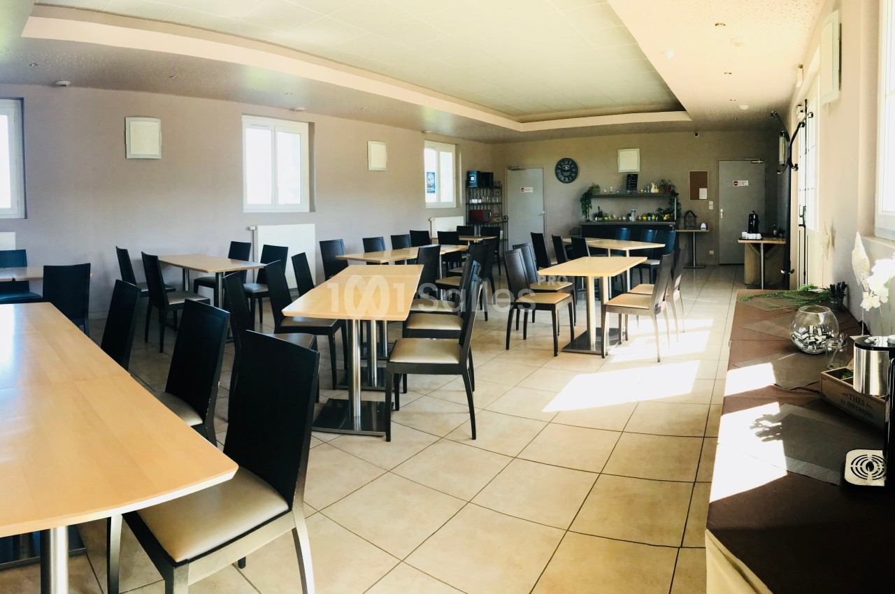 Salle lumineuse avec tables en bois, chaises noires, carrelage au sol et coin buffet en arrière-plan.