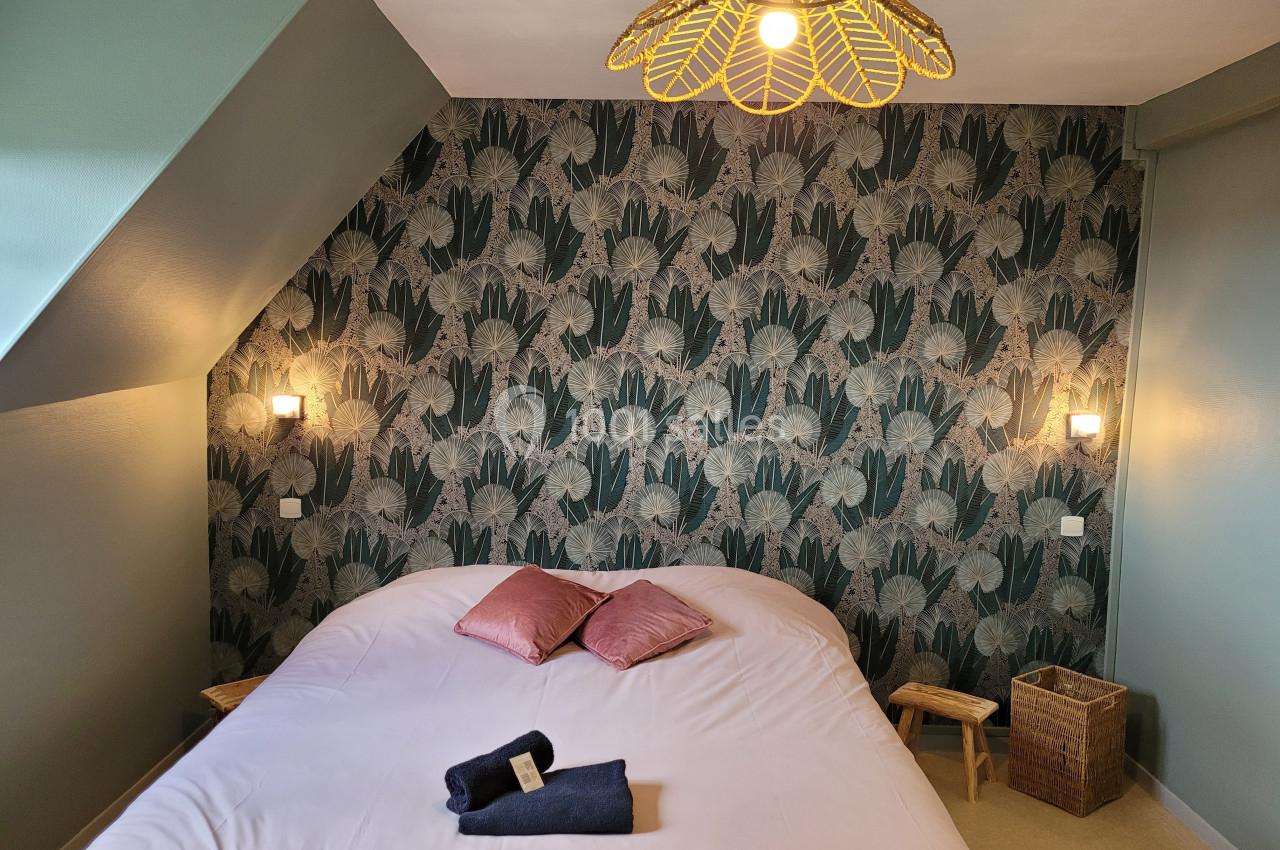 Chambre avec lit double, mur tapissé de motifs végétaux, éclairage mural et suspension en osier.