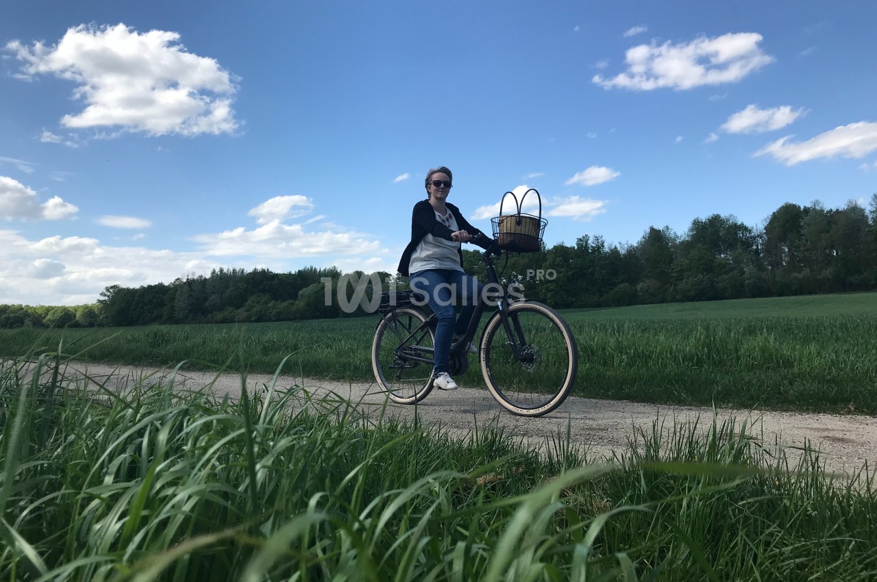 Une personne à vélo sur un chemin de campagne bordé de verdure, sous un ciel bleu avec quelques nuages.