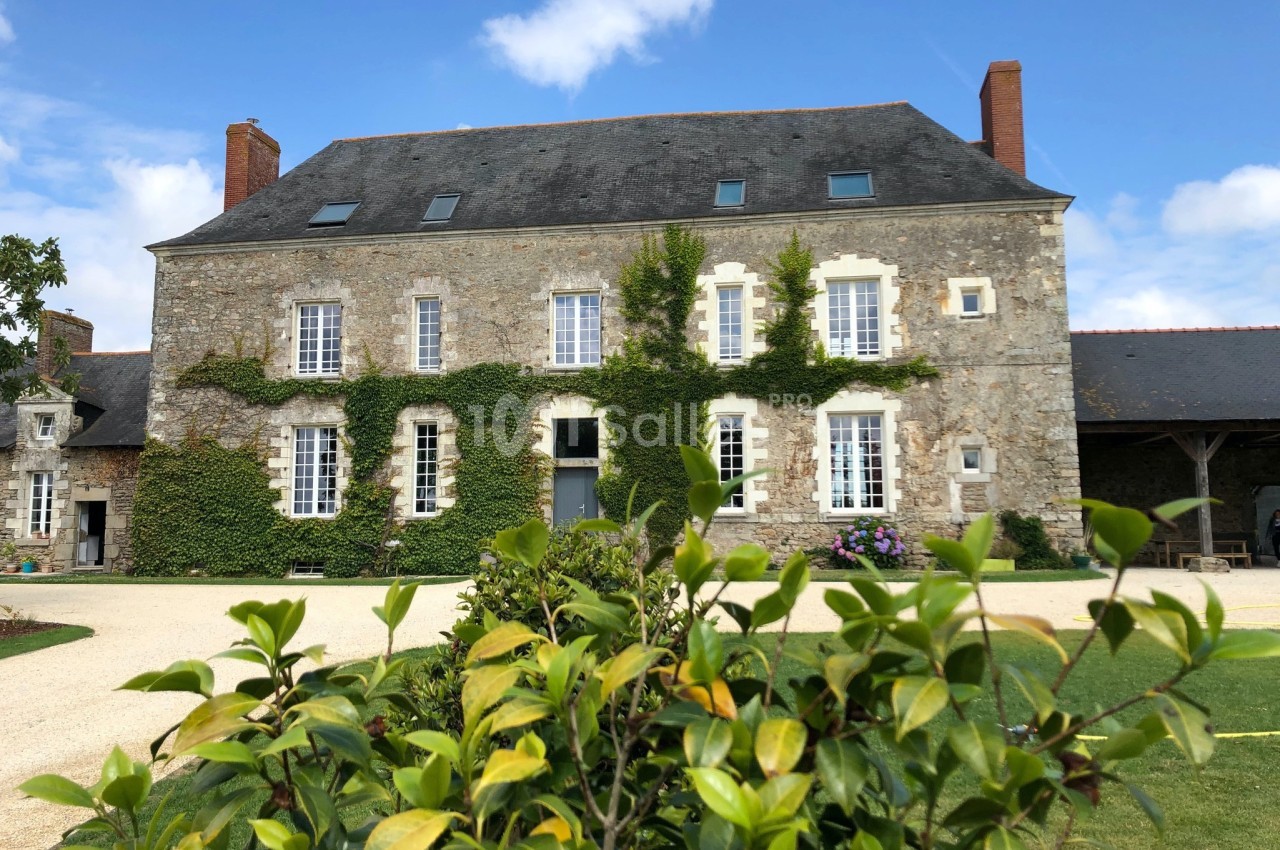 Façade d'une maison en pierre avec des volets blancs, entourée de verdure et d'un jardin sous un ciel bleu.