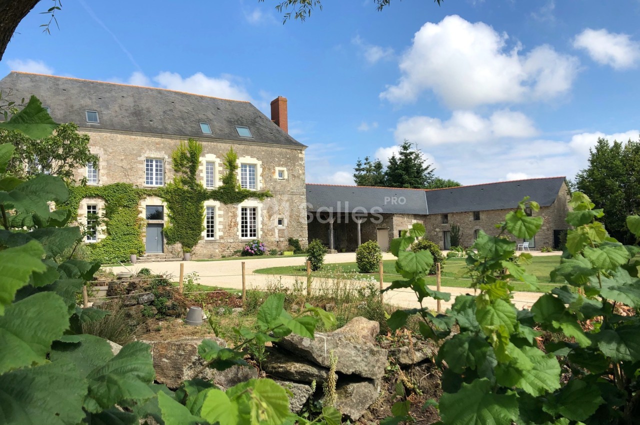 Bâtisse en pierre avec volets bleus, entourée de verdure et d'une cour, sous un ciel partiellement nuageux.