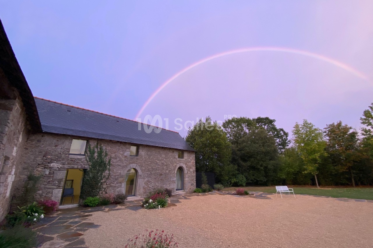 Bâtisse en pierre avec toit en ardoise, cour gravillonnée, végétation et arc-en-ciel dans un ciel pastel.