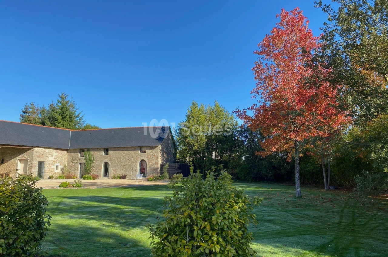 Cour verdoyante avec pelouse, arbres aux feuilles automnales et bâtiment en pierre sous un ciel bleu clair.