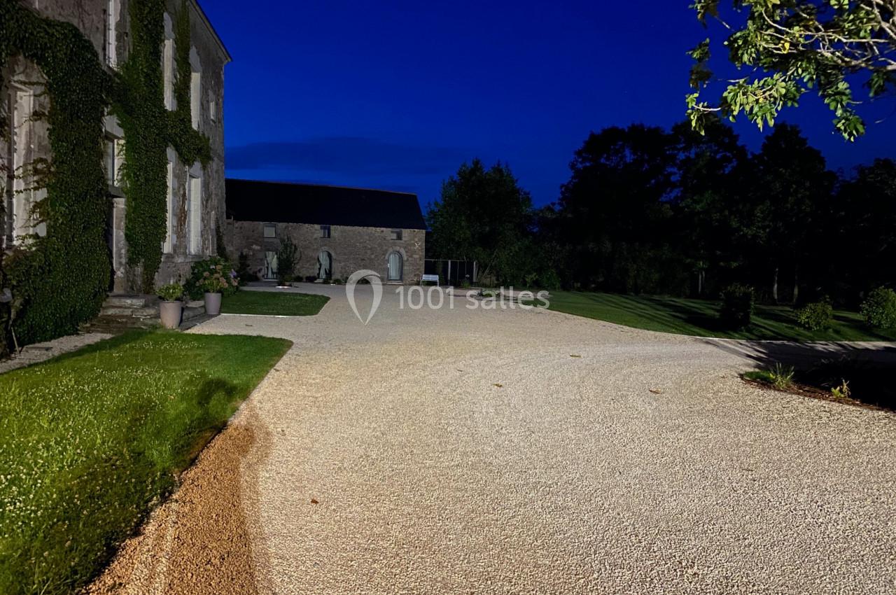 Allée en gravier éclairée la nuit, bordée de pelouses et d'un bâtiment en pierre recouvert de végétation.