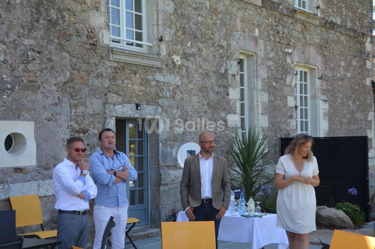 Quatre personnes discutent devant un bâtiment en pierre, avec des chaises jaunes et une table dressée à proximité.