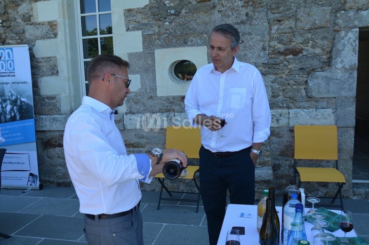 Deux hommes en chemise discutent près d'une table avec des bouteilles de vin et de l'eau, devant un mur en pierre.