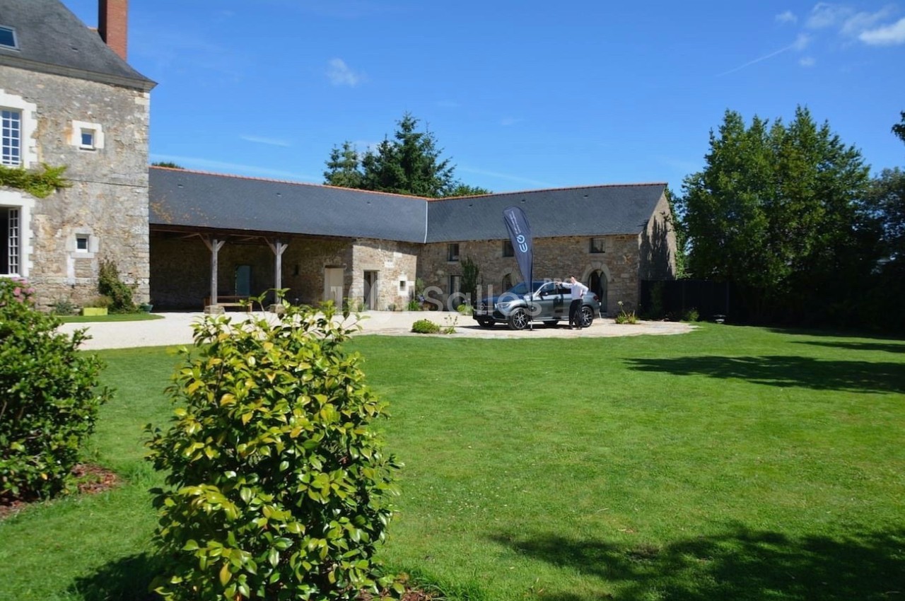 Cour verdoyante avec une bâtisse en pierre, une voiture stationnée près d'un drapeau et un ciel dégagé.