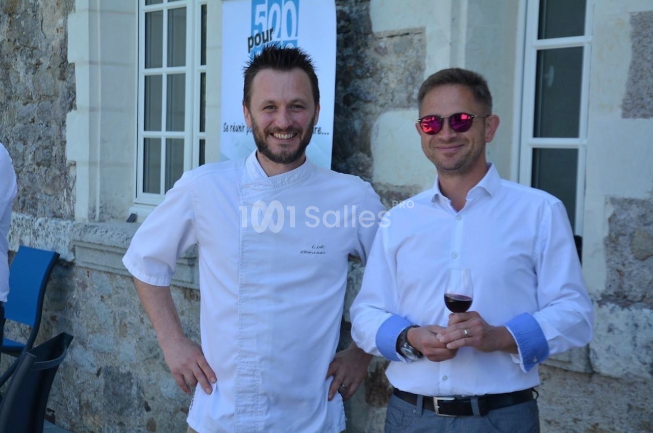 Deux hommes souriants, l'un en tenue de chef, l'autre en chemise blanche tenant un verre de vin, devant un bâtiment en…