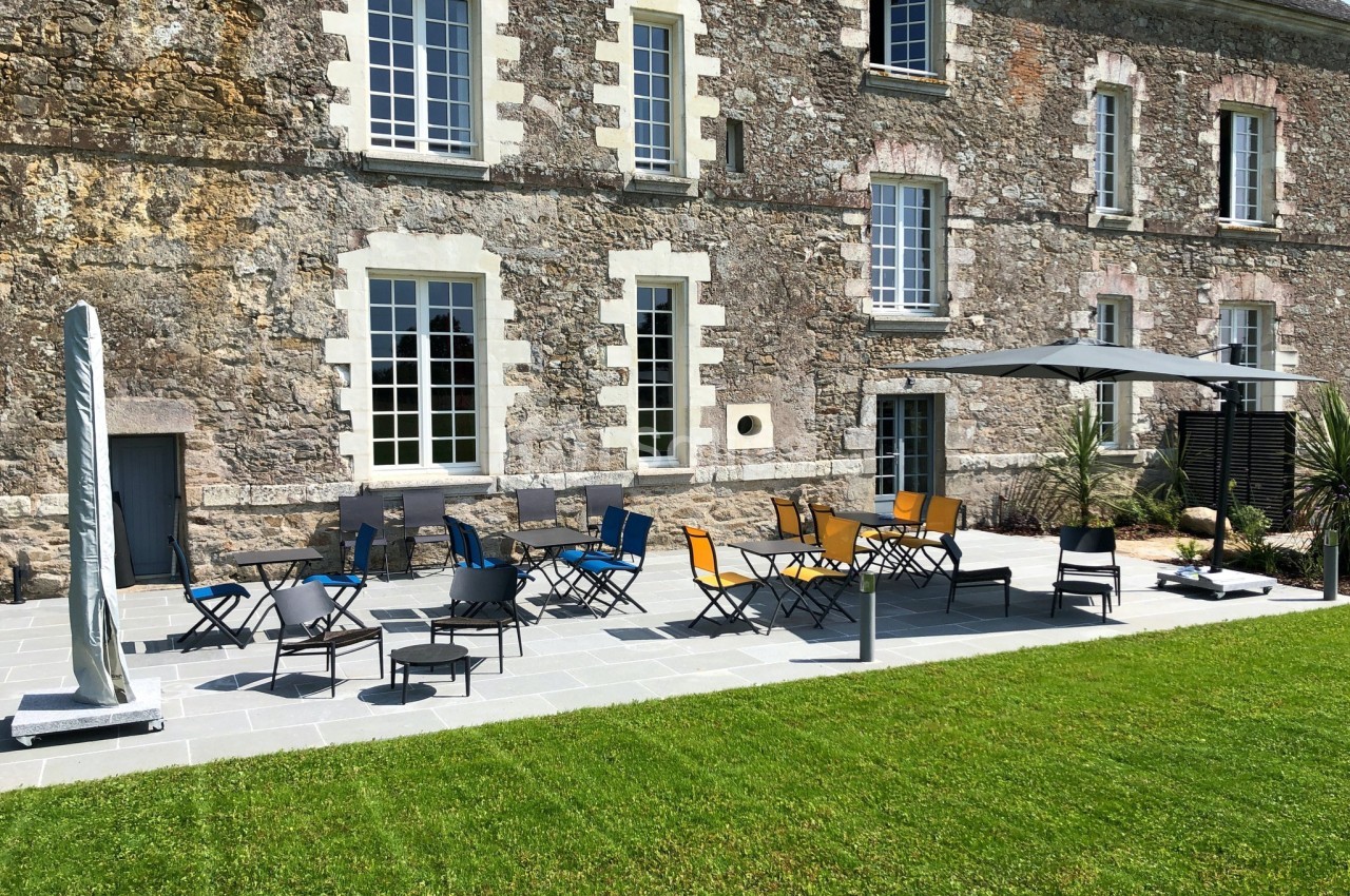 Terrasse en pierre avec tables, chaises colorées et parasol, devant une façade en pierre avec grandes fenêtres.