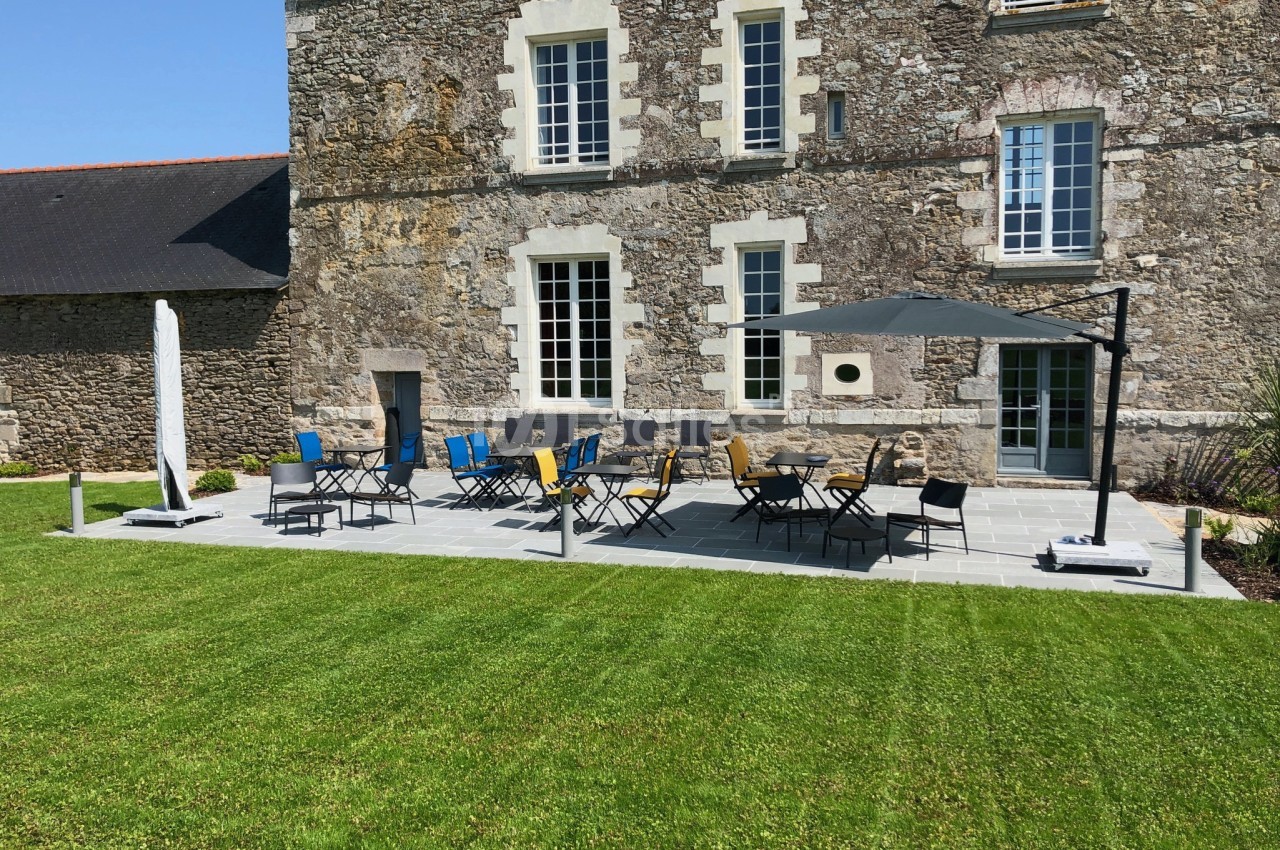 Terrasse en pierre avec tables, chaises colorées et parasols, devant une bâtisse en pierre entourée de pelouse.