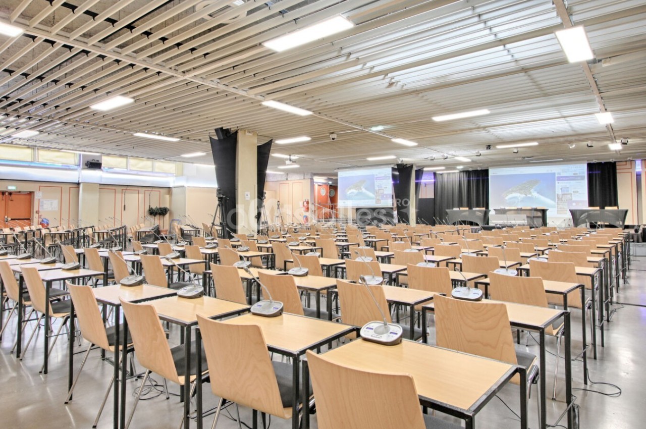 Salle de conférence moderne avec rangées de tables et chaises, équipée de micros et écrans de projection.