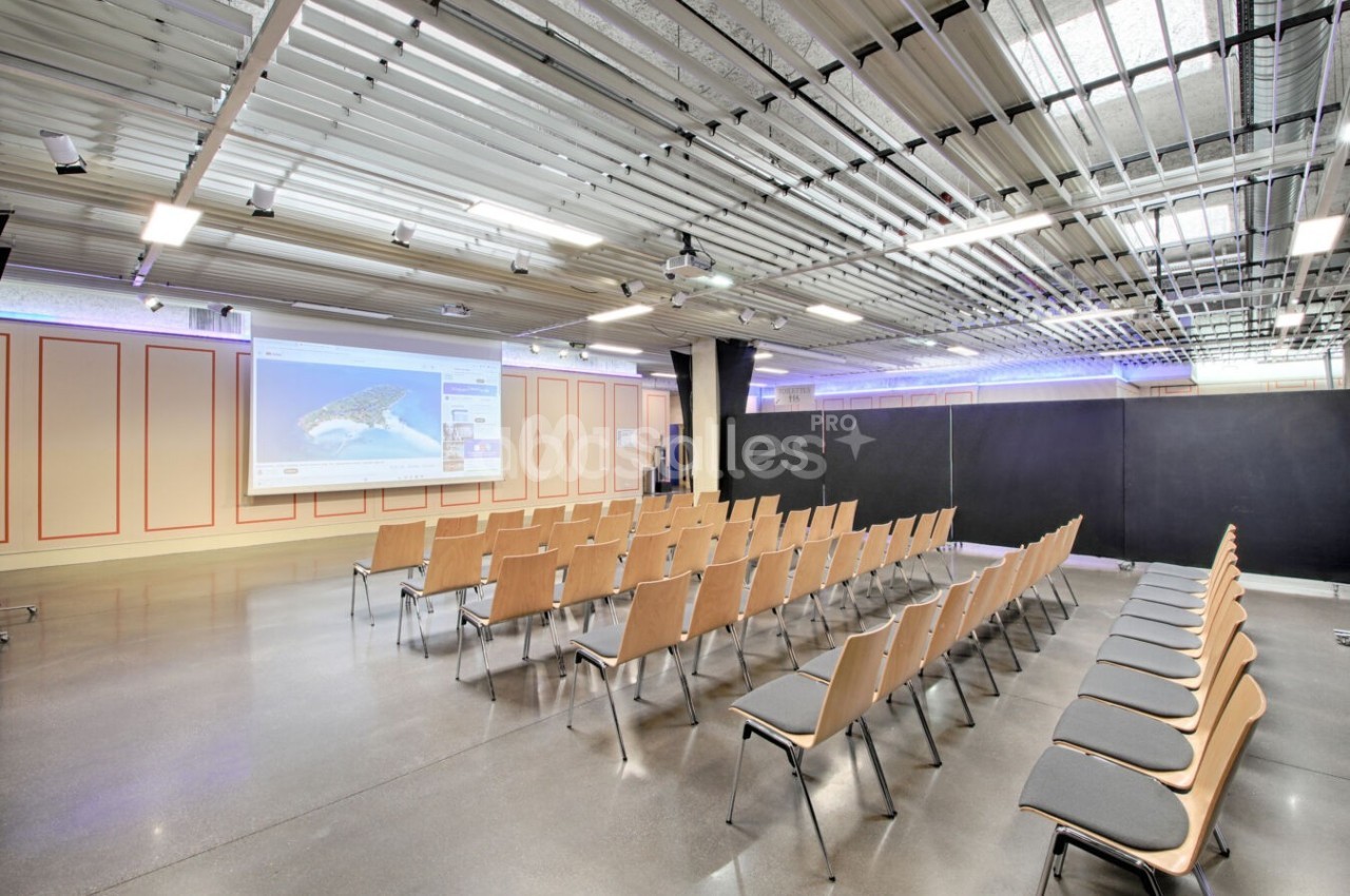 Salle de conférence moderne avec rangées de chaises en bois, écran de projection allumé et plafond industriel visible.