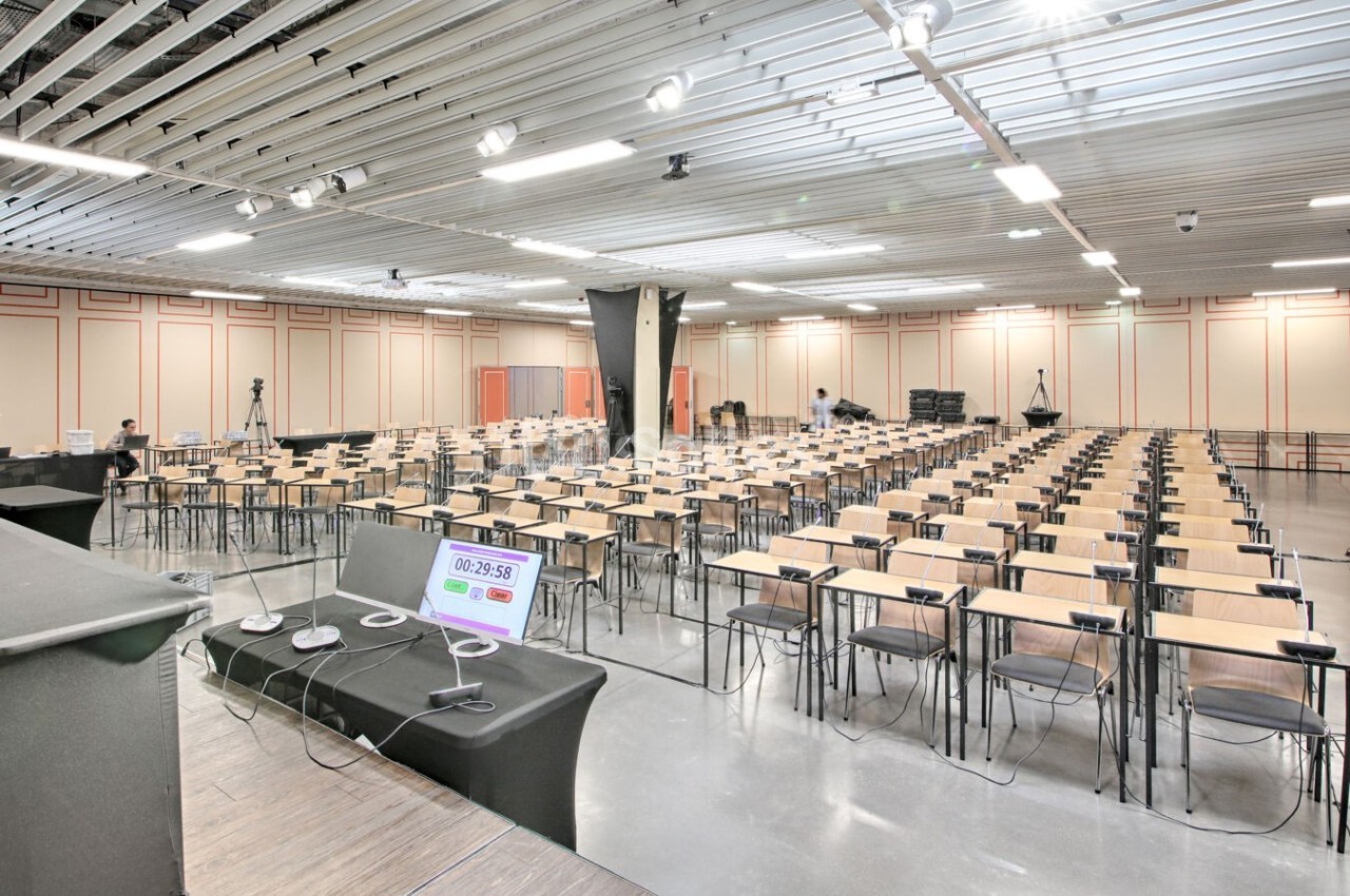 Salle de conférence vide avec des rangées de tables et chaises, vue depuis une estrade équipée de micros et d'un écran.