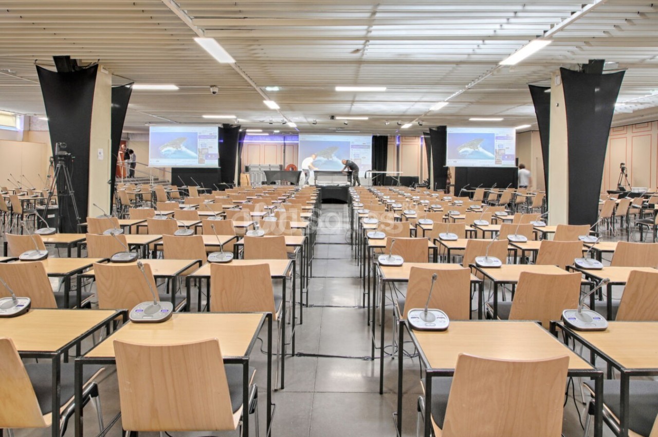 Salle de conférence équipée de rangées de tables, chaises et micros, avec écrans et projecteurs en arrière-plan.