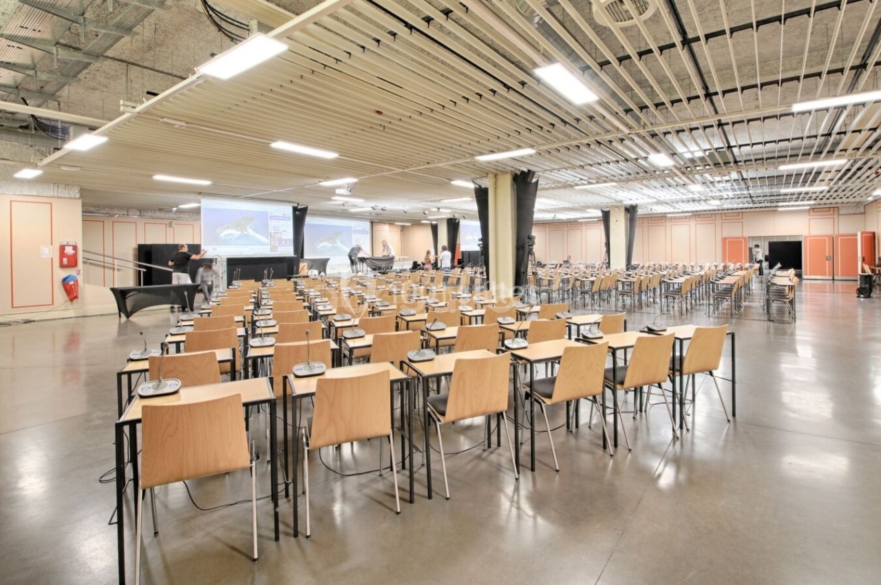 Salle de conférence spacieuse avec des rangées de chaises en bois et des tables, équipée de projecteurs et d'écrans.