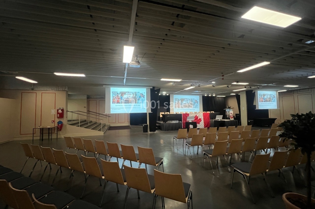 Salle de conférence vide avec des rangées de chaises, un podium, des écrans projetant des présentations et un drapeau…