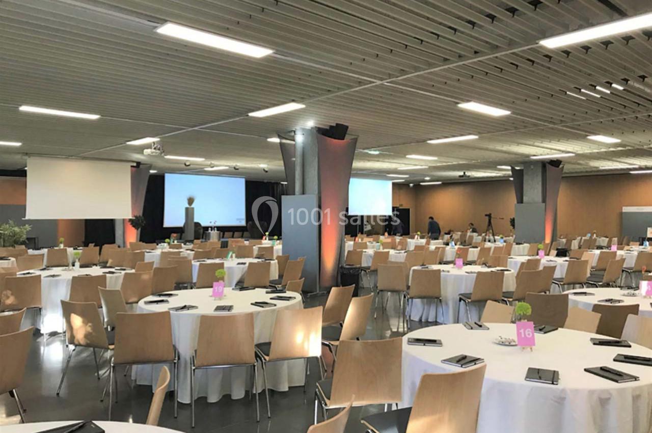Salle de conférence aménagée avec des tables rondes, des chaises en bois et des écrans de projection en arrière-plan.