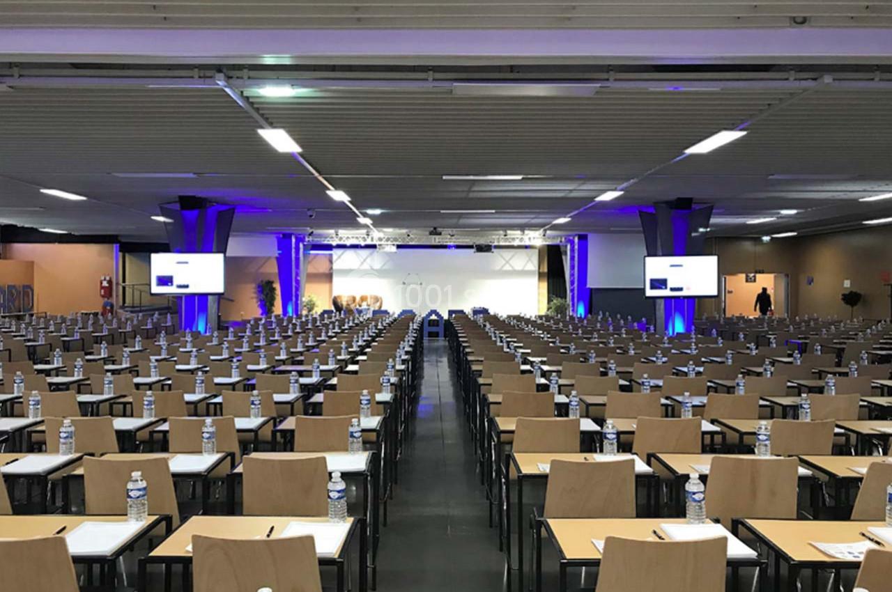 Salle de conférence avec de nombreuses rangées de tables et chaises, équipée d'écrans et de bouteilles d'eau sur chaque…