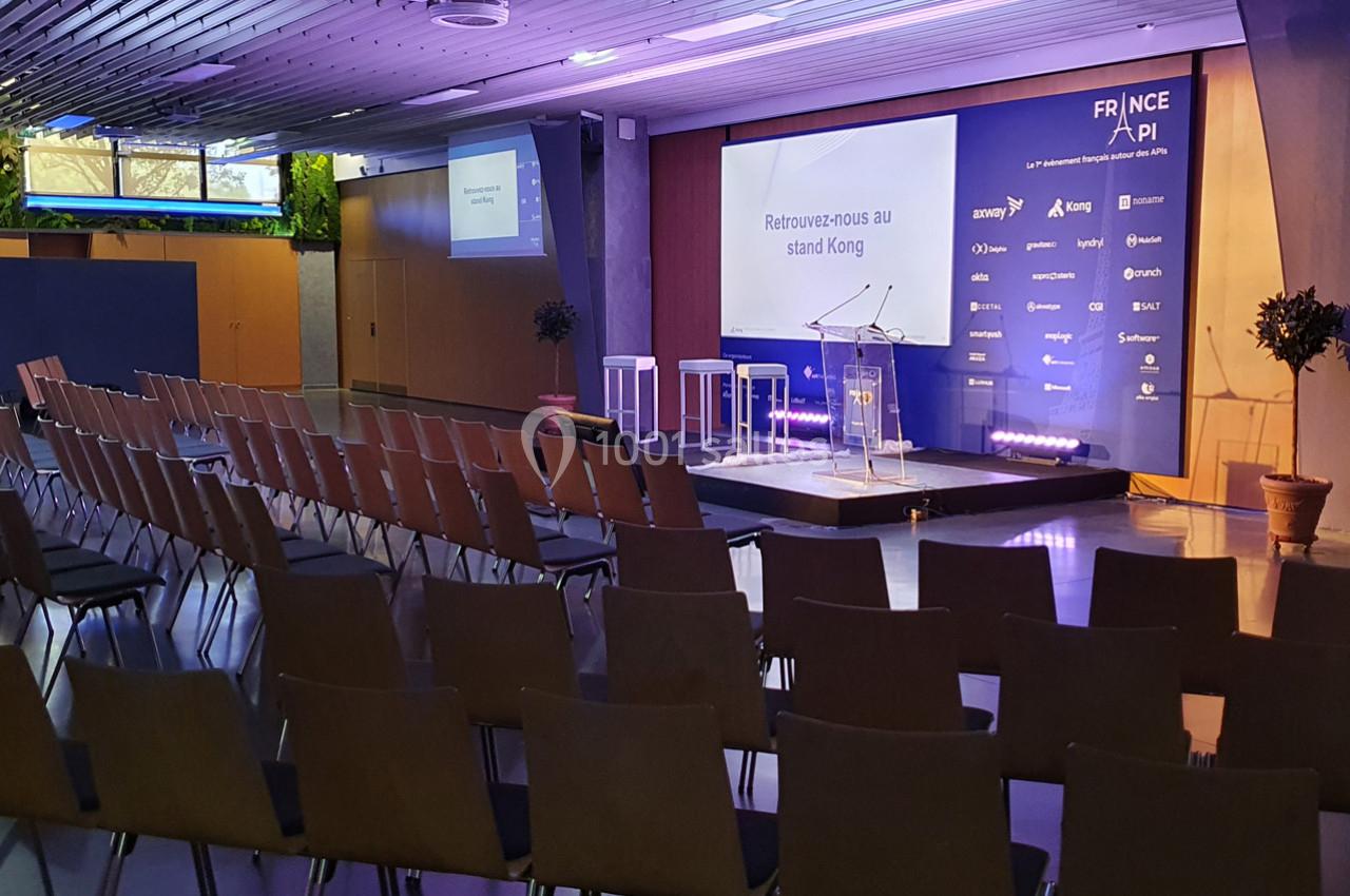 Salle de conférence vide avec des rangées de chaises et une scène équipée d'un pupitre, éclairée par des projecteurs.