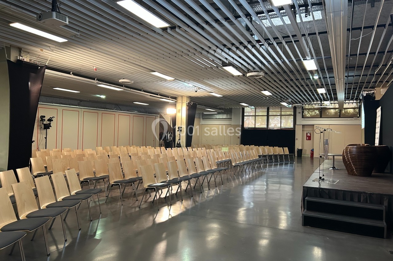 Salle de conférence vide avec des rangées de chaises en bois, une estrade et des équipements audiovisuels.